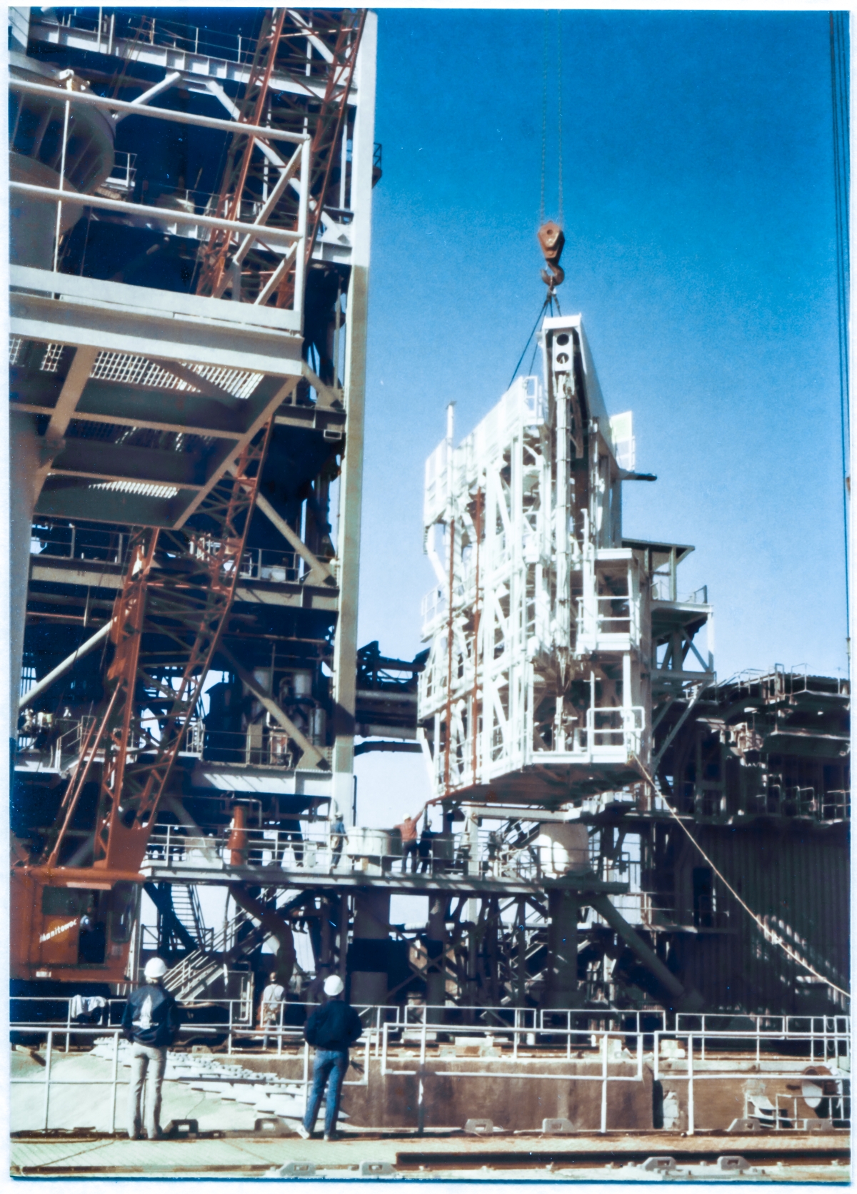 Image 122. At Space Shuttle Launch Complex 39-B, Kennedy Space Center, the IAA (Intertank Access Arm) no longer rests on its special transporter and is now in free suspension above the Pad Deck. Union Ironworkers from Local 808, working for Ivey Steel Erectors, can be seen on the MLP Utilities Interface Platform, across the Flame Trench, still hands-on with it (barely), continuing to guide and orient it in conjunction with another (unseen) ironworker who is holding the tagline attached to the IAA's lower right corner, which extends down and to the right, across the darkness of the 9099 Building, out of frame. The IAA weighs roughly 100,000 pounds and working it into position is neither easy nor safe to do, but it must be done, and Union Ironworkers are up to the task. The IAA will be bolted to the Fixed Service Structure, which dominates the left side of this image, in the orientation you see it here, between the 200'-0” and 220'-0” levels, protruding outward toward the east from where it will be attached to its Support Structure which runs along Side 4 (the north side) of the FSS. Photo by James MacLaren.